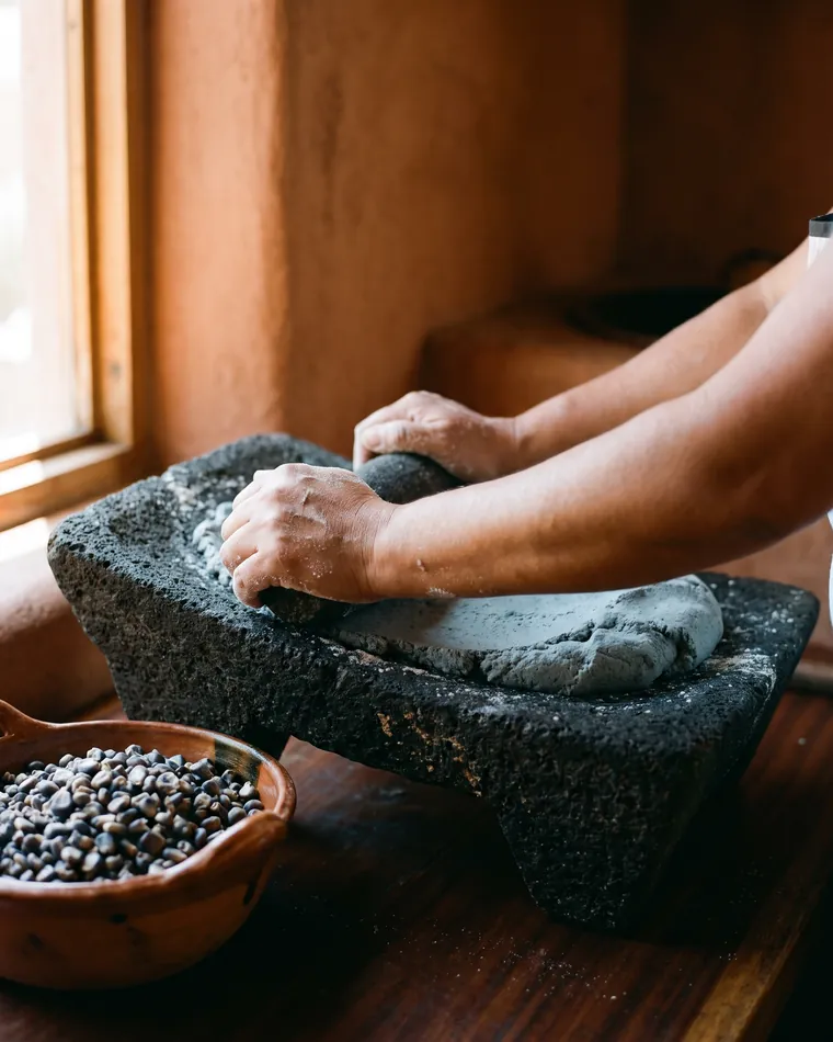 Unas manos muelen el nixtamal azul en un metate de piedra volcánica; la masa se junta al frente.