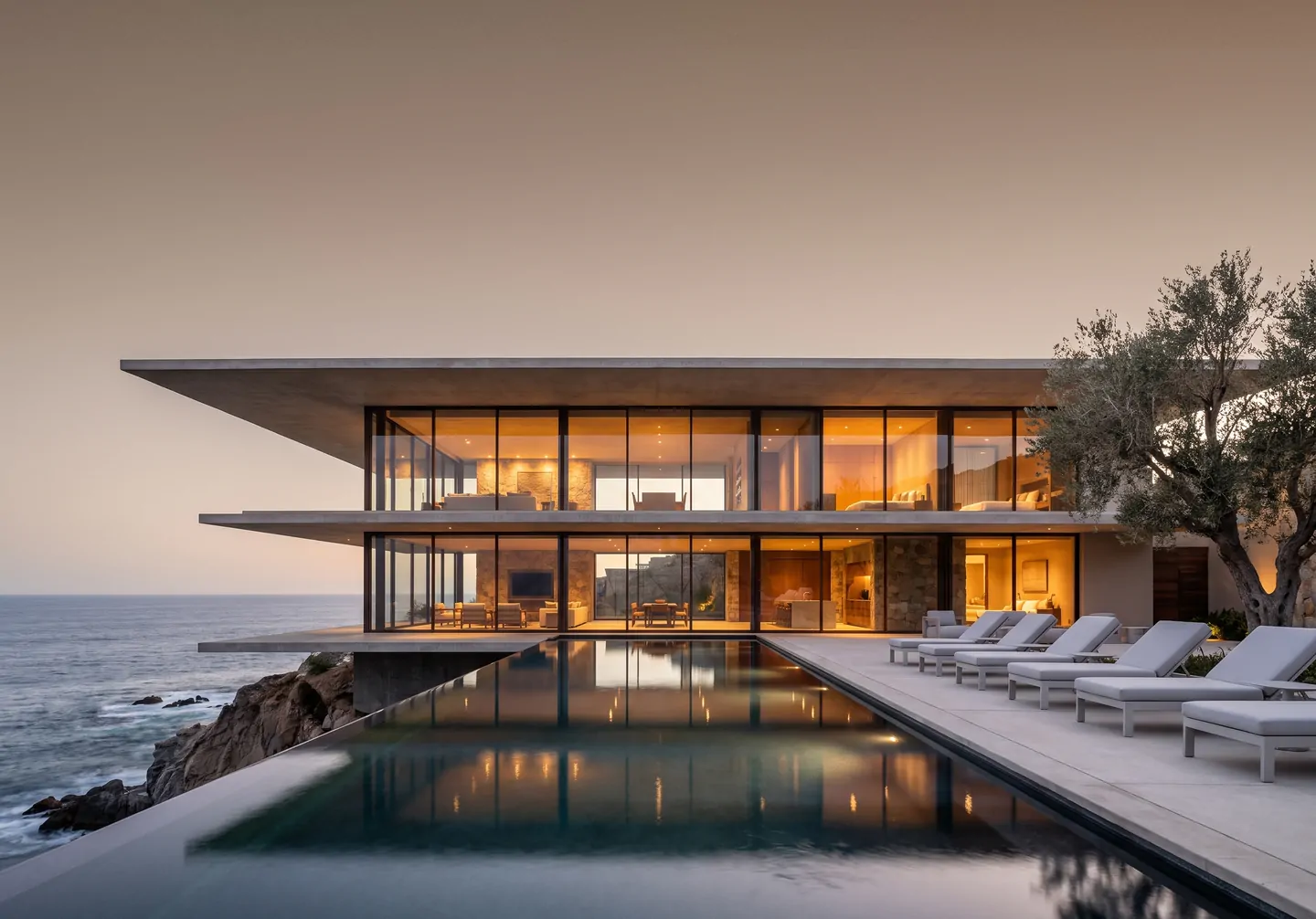 Casa Acantilado al atardecer: residencia de concreto con techo volado, muros de vidrio iluminados, un olivo, camastros y alberca infinita sobre el acantilado frente al océano Pacífico