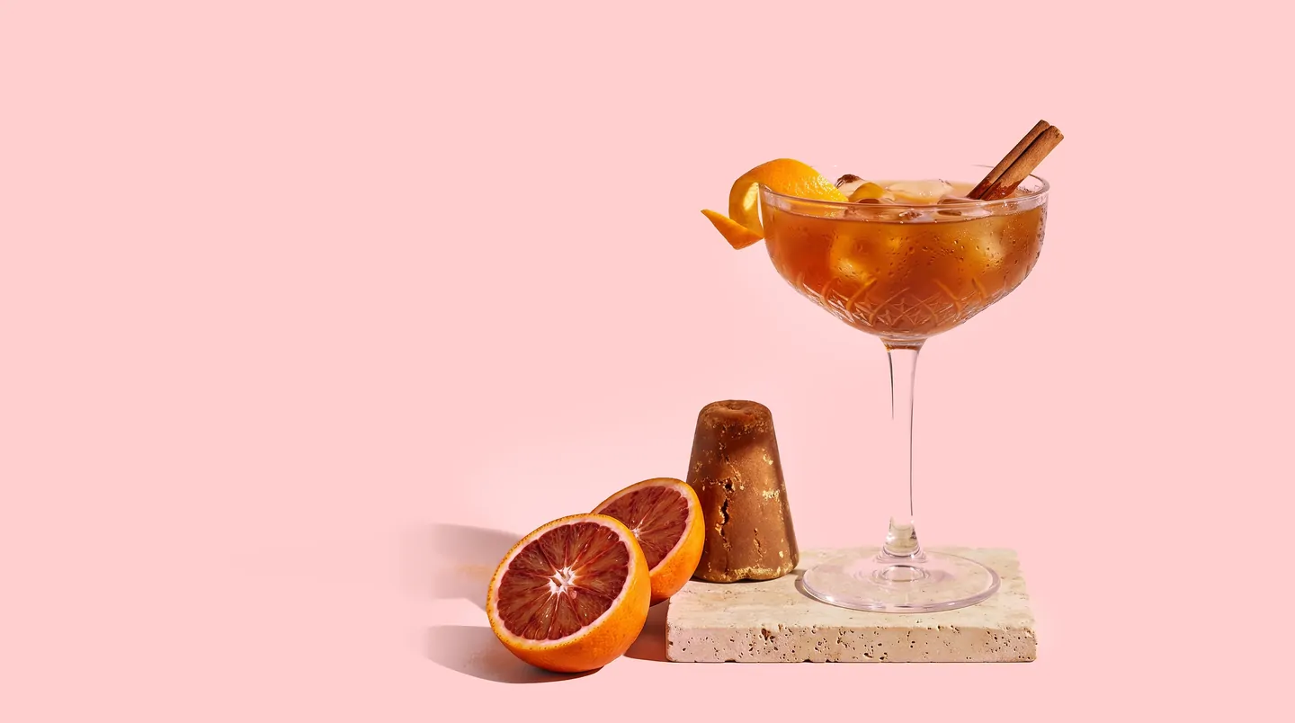 Copa de cristal tallado con bebida ámbar, piel de naranja y canela, junto a naranjas sanguinas y un cono de piloncillo sobre fondo rosa